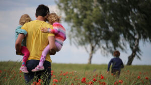 A person carrying two children through a field as another child runs ahead of them.