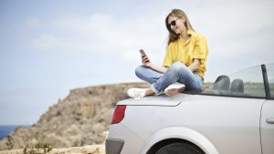 A person sits on the trunk of a rental car as they use their cell phone.